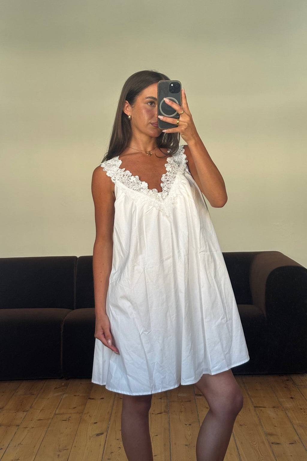 Woman taking a mirror selfie wearing a white dress in a room with a black couch.