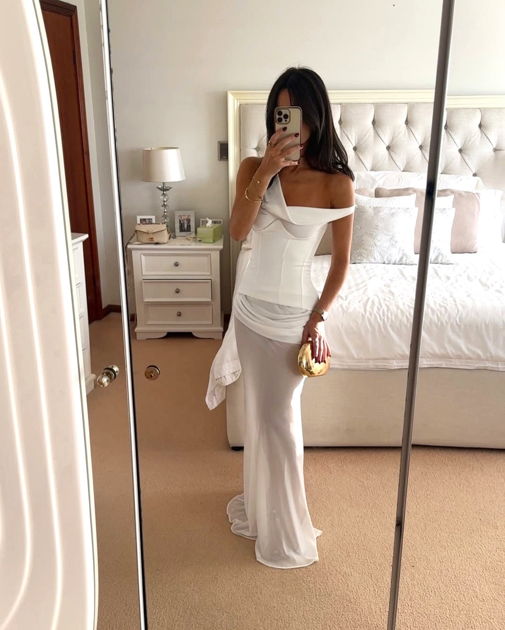 Woman in a white dress taking a mirror selfie in a bedroom.
